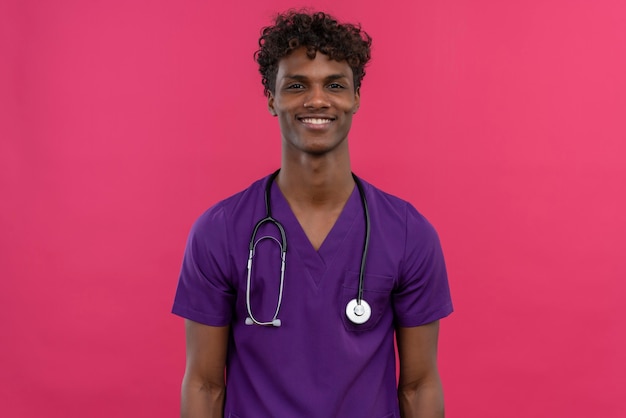 Szczęśliwy młody przystojny ciemnoskóry lekarz z kręconymi włosami ubrany w fioletowy mundur ze stetoskopem uśmiechnięty, podczas gdy