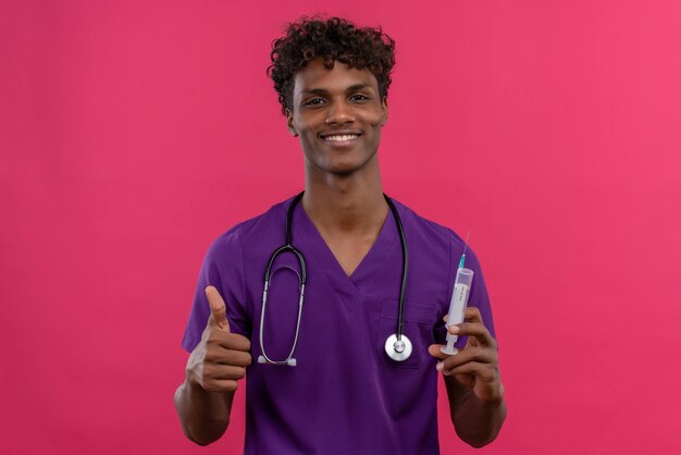 Szczęśliwy młody przystojny ciemnoskóry lekarz z kręconymi włosami w fioletowym mundurze ze stetoskopem pokazującym kciuki do góry, trzymając strzykawkę do wstrzyknięć
