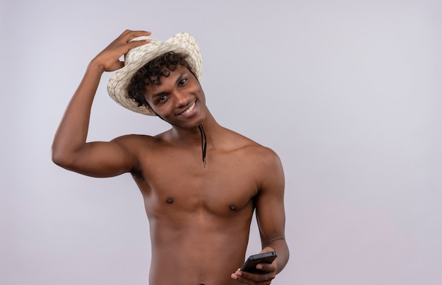 Szczęśliwy młody przystojny ciemnoskóry mężczyzna z kręconymi włosami na sobie kapelusz przeciwsłoneczny, uśmiechając się i trzymając telefon komórkowy