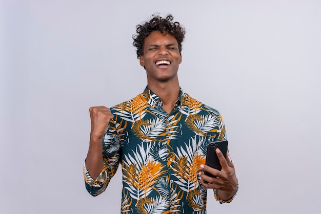 Szczęśliwy młody przystojny ciemnoskóry mężczyzna z kręconymi włosami w koszuli z nadrukiem liści, trzymając smartfon z zaciśniętą pięścią