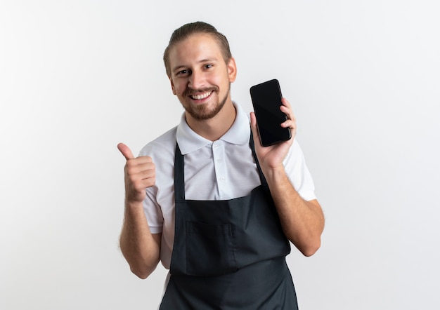 Szczęśliwy młody przystojny fryzjer na sobie mundur pokazujący telefon komórkowy i kciuk na białym tle na biały z miejsca na kopię