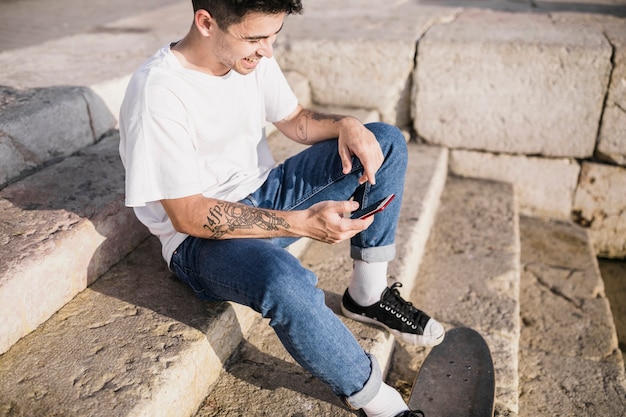 Szczęśliwy nastolatek siedzi na schody z deskorolka i telefon komórkowy
