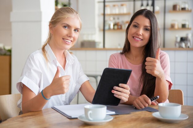 Szczęśliwy, odnoszący sukcesy agent i zadowolony klient, pokazując kciuk w górę, siedząc przy stole i razem używając tabletu