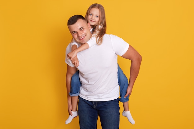 szczęśliwy ojciec i mała dziewczynka w białych t-shirtach i dżinsach, pozowanie na żółtym, mają szczęśliwy wyraz twarzy, spędzają razem czas. Koncepcja rodziny.