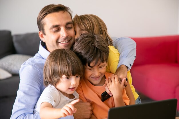 Szczęśliwy ojciec przytulanie z uroczymi dziećmi. Kaukaski tata w średnim wieku siedzi w salonie, obejmując słodkie dzieci, trzymając telefon komórkowy i uśmiechając się. Koncepcja ojcostwa, dzieciństwa i rodziny