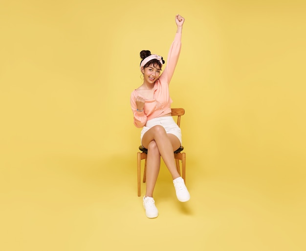 Szczęśliwy piękny Asian teen kobieta siedzi na krześle i ręce w górę docenianie gest na żółtej ścianie. Koncepcja sukcesu podekscytowany zwycięzca.