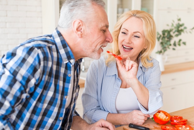 Bezpłatne zdjęcie szczęśliwy portret starsza kobieta karmi plasterek dzwonkowy pieprz jej mąż w kuchni
