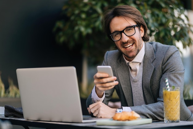 Szczęśliwy przedsiębiorca wysyła SMS-y na telefon komórkowy, siedząc na przerwie w kawiarni na świeżym powietrzu