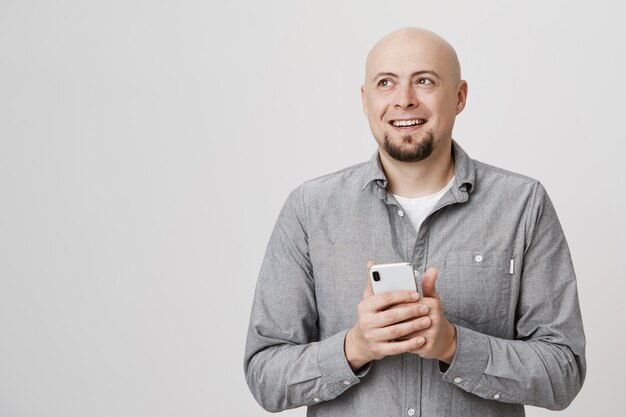 Szczęśliwy przemyślany łysy mężczyzna odwrócić wzrok, uśmiechając się jak przy użyciu telefonu komórkowego