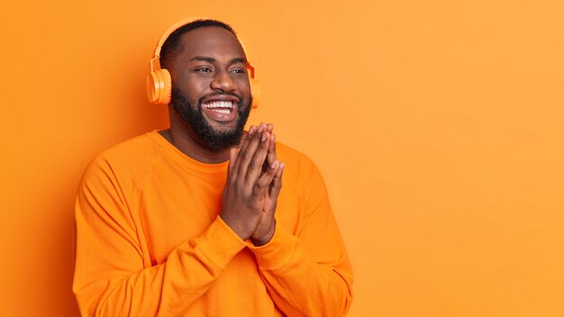 Szczęśliwy pulchny mężczyzna trzyma dłonie przyciśnięte do siebie, uśmiecha się szeroko ubrany w swobodny sweter, nosi słuchawki stereo, słucha przyjemnych melodii na pomarańczowej ścianie studia z miejscem na kopię