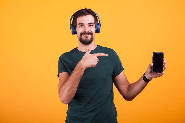 Szczęśliwy radosny człowiek nosi słuchawki i wskazuje na swojego smartfona na białym tle na pomarańczowym tle w studio.