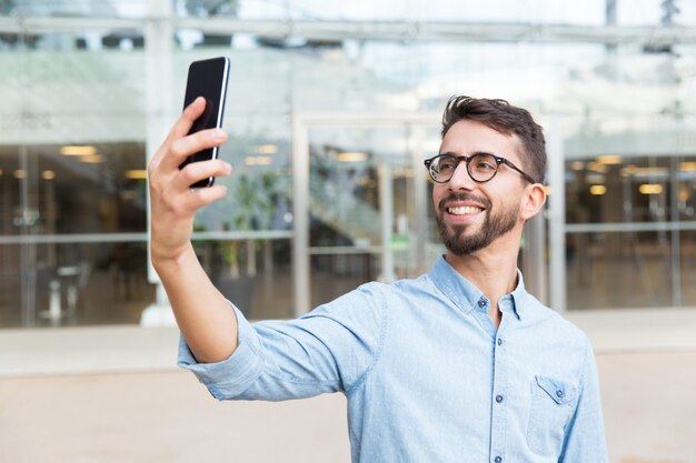 Szczęśliwy radosny facet w okularach biorąc selfie na smartfonie