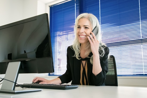 Bezpłatne zdjęcie szczęśliwy siwy bizneswoman rozmawia z kolegą na telefon. profesjonalny menedżer pewny siebie rozmawiający, uśmiechnięty i pracujący na komputerze. koncepcja biznesowa, miejsce pracy i komunikacji