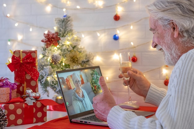 Szczęśliwy starszy mężczyzna w rozmowie wideo z laptopem świętuje boże narodzenie przy lampce wina musującego. szczęśliwego nowego roku i wesołych świąt