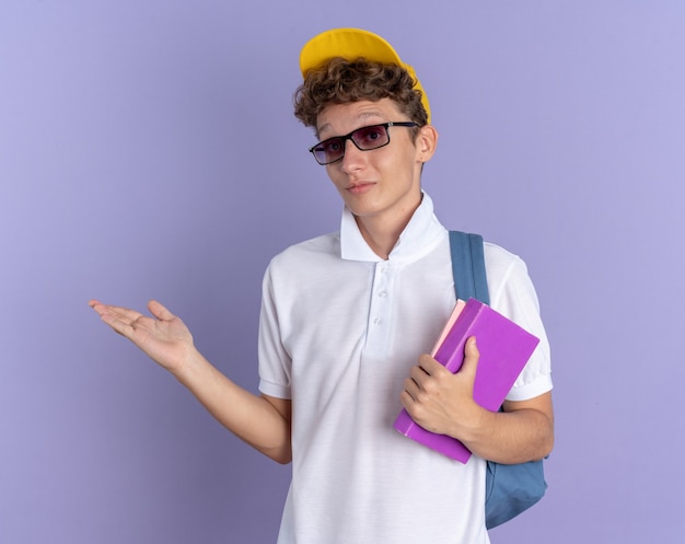 Szczęśliwy student facet w białej koszulce polo i żółtej czapce w okularach z plecakiem, trzymając zeszyty, patrząc na kamerę, uśmiechając się, prezentując ręką stojącą na niebieskim tle