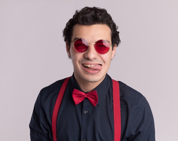 Szczęśliwy stylowy mężczyzna z muszką w okularach i szelkach, patrząc na przód wystający język stojący na białej ścianie