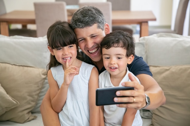 Szczęśliwy tata i dwoje dzieci, biorąc selfie lub rozmawiając przez telefon, siedząc razem na kanapie w domu.