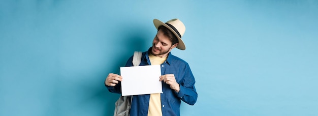 Bezpłatne zdjęcie szczęśliwy turysta w słomkowym kapeluszu uśmiechający się i patrzący na kartkę papieru dla twojego logo stojącego na niebieskim grzbiecie