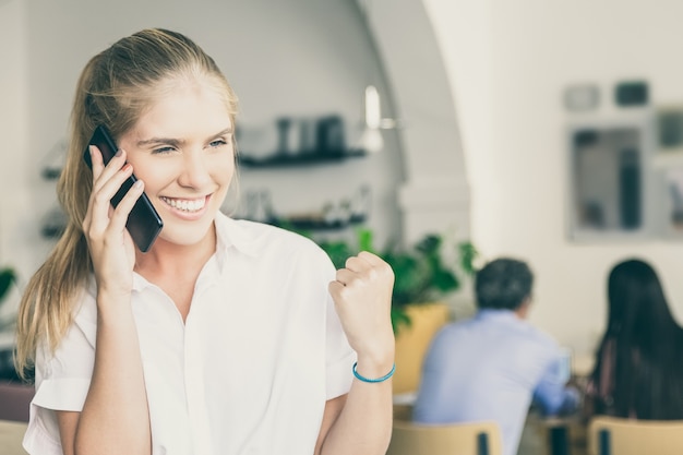 Szczęśliwy udany piękna młoda kobieta rozmawia przez telefon komórkowy, czyniąc gest zwycięzcy, stojąc w przestrzeni coworkingowej