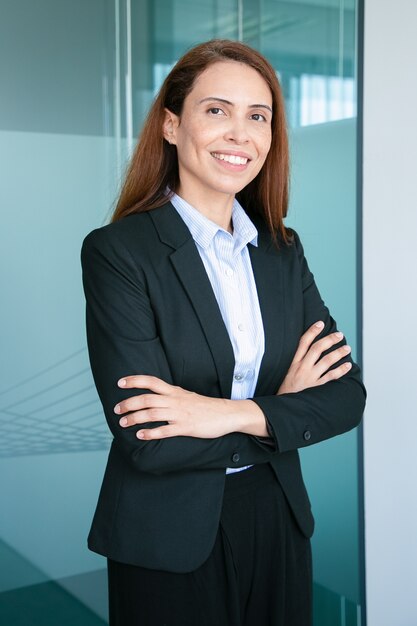 Szczęśliwy udany rudowłosy bizneswoman noszenie formalnego garnituru, stojąc z założonymi rękoma i uśmiechnięty