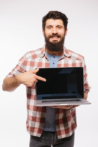Szczęśliwy uśmiechnięty brodaty mężczyzna trzyma pusty ekran laptopa na białym tle nad białą ścianą