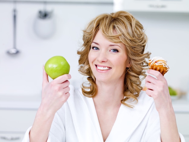 Bezpłatne zdjęcie szczęśliwy wesoły młoda piękna kobieta z zielonym jabłkiem i słodkim ciastem siedzi w kuchni