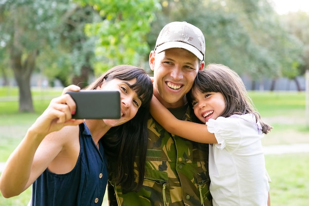 Szczęśliwy wojskowy, jego żona i córeczka, biorąc selfie na telefon komórkowy w parku miejskim. Przedni widok. Zjazd rodzinny lub koncepcja powrotu do domu