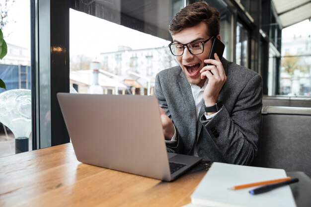 Szczęśliwy zdziwiony biznesmen siedzi stołem w kawiarni w eyeglasses podczas gdy opowiadający smartphone i używać laptop