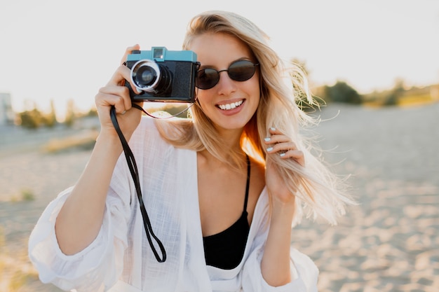Bezpłatne zdjęcie szczupła blond szczęśliwa kobieta trzyma aparat retro i zabawę na ciepłej słonecznej plaży. koncepcja wakacji i podróży. naturalne piękno, wakacje w azji. modne okulary przeciwsłoneczne, biały strój.