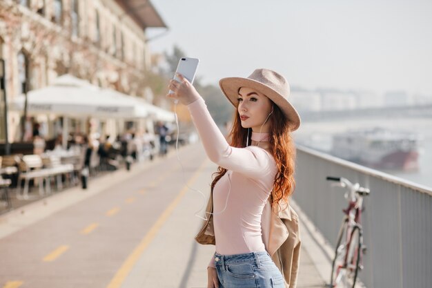 Szczupła kobieta w modnym beżowym kapeluszu robi selfie z poważnym wyrazem twarzy