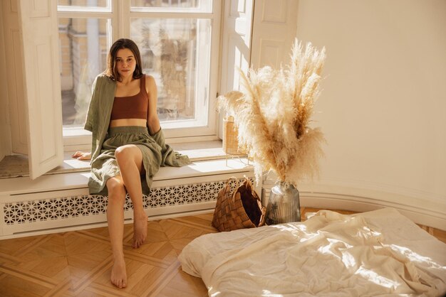Szczupła młoda dziewczyna kaukaski patrzy na kamerę siedząc na parapecie przy oknie we wnętrzu pokoju Brunetka ubrana jest w koszulkę i szorty latem Relaks w domu w pomieszczeniu