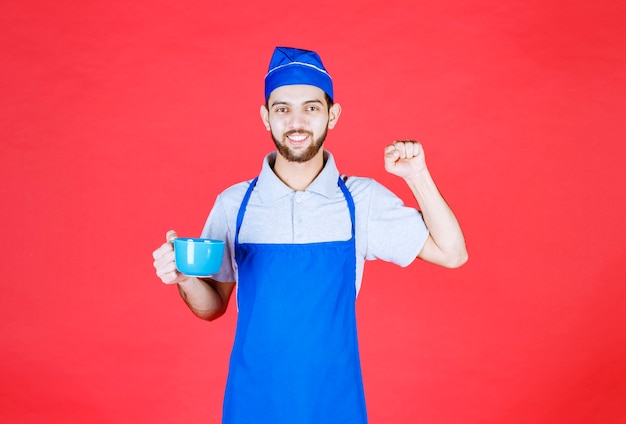 Szef kuchni w niebieskim fartuchu trzymający niebieski ceramiczny kubek i pokazujący pięść.