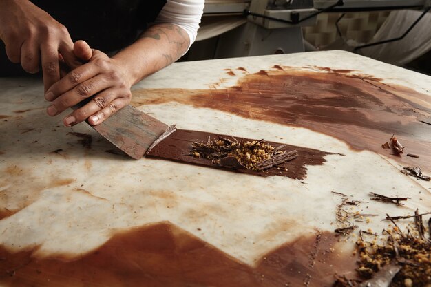 Szef Murzyn zbiera schłodzoną roztopioną czekoladę z marmurowego stołu, abstrakcyjny obraz zbliżenia pracy w cukierni czekoladowej