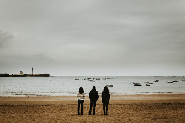 Szerokie ujęcie trzech osób stojących w pobliżu morza z małymi łódkami unoszącymi się w morzu