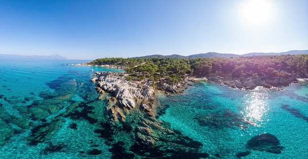 Szerokie ujęcie wybrzeża Morza Egejskiego z niebieską przezroczystą wodą, zielenią dookoła, skałami, krzewami i drzewami, wzgórzami, widokiem na pamoramę z drona, Grecja