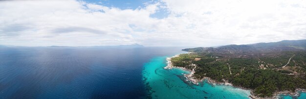 Szerokokątne ujęcie skalistego wybrzeża Morza Egejskiego z zielenią wokół, krzewami i drzewami, wzgórzami i górami, błękitną wodą z falami, widok z drona Grecja