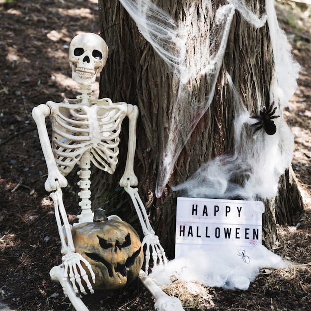 Szkielet siedzi w pobliżu drzewa z dyni i Happy Halloween napis