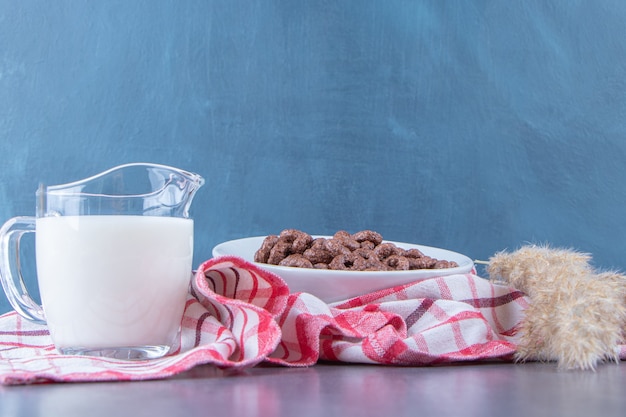 Szklanka mleka obok krążków kukurydzy w szklanej misce obok trawy pampasowej na ściereczce, na marmurowym stole.