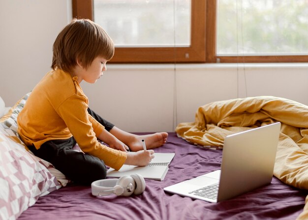 Szkoła chłopiec siedzi w łóżku z laptopem i słuchawkami
