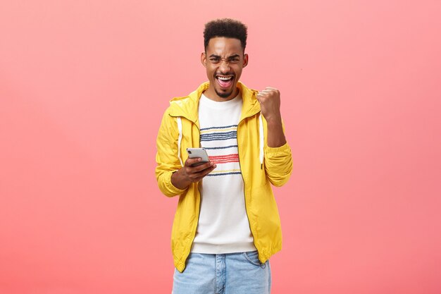 Tak, wygrałem. Portret szczęśliwego podekscytowanego i usatysfakcjonowanego afroamerykańskiego mężczyzny z brodą podnoszącą pięść w geście zwycięstwa i triumfu, cieszący się, trzymając smartfon wygrywający w grze telefonicznej online nad różową ścianą