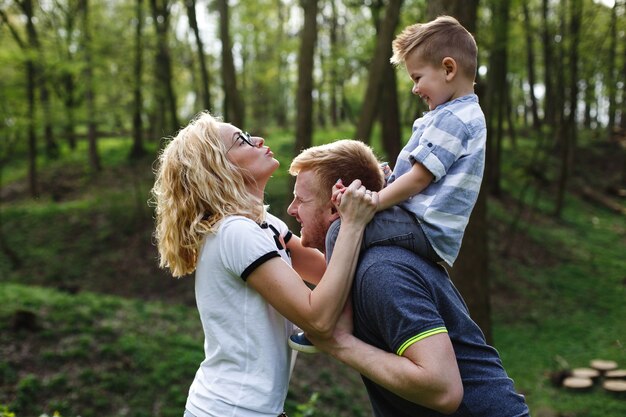 Tata trzyma syna na karku i bawi się z mamą w zielonym letnim parku