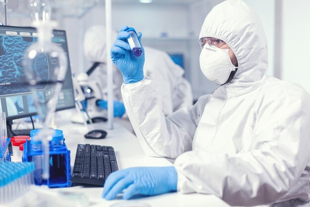 Technik laboratoryjny ubrany w kombinezon ochronny jako środek ostrożności, patrząc na probówkę