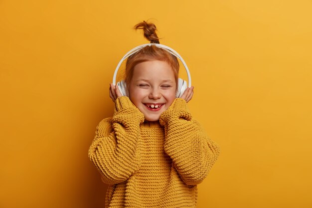 Technologia, dzieci, koncepcja muzyki. Całkiem uśmiechnięte małe dziecko z rudymi włosami nosi słuchawki stereo, cieszy się czystym dźwiękiem i słucha ulubionej piosenki, radośnie chichocze, nosi sweter z dzianiny
