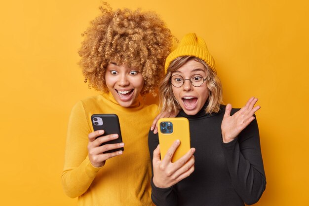 Technologia i koncepcja przyjaźni Szalone, pozytywne kobiety używają telefonów komórkowych, patrzą na ekran, reagują na niesamowite wiadomości, ubierają się w luźne golfy odizolowane nad żółtą ścianą, zauważają coś niewiarygodnego