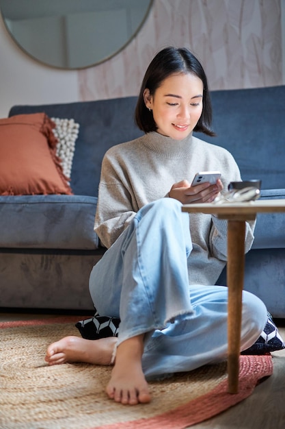 Technologia i ludzie młoda stylowa azjatycka kobieta siedzi w domu ze swoim smartfonem wysyłając wiadomość tekstową usi