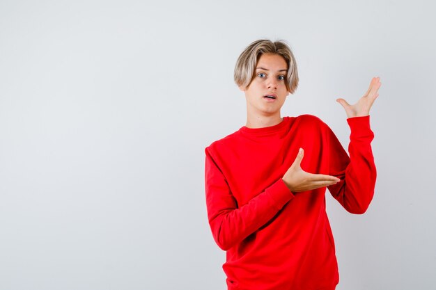 Teen chłopiec udając, że pokazuje coś w czerwonym swetrze i patrząc zdziwiony, widok z przodu.