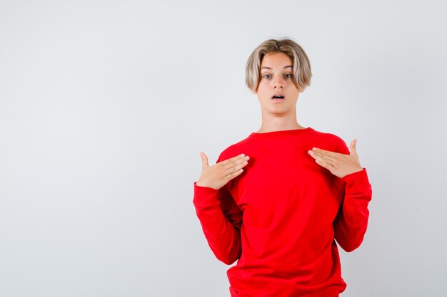 Teen chłopiec w czerwonym swetrze z rękami na klatce piersiowej i patrząc zdumiony, widok z przodu.