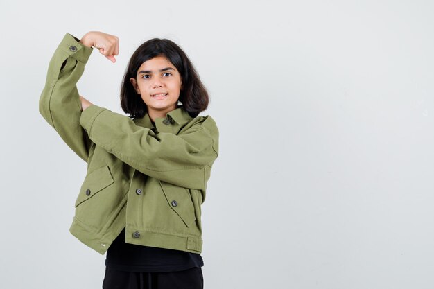 Teen dziewczyna pokazuje mięśnie ramion w t-shirt, zieloną kurtkę i patrząc wesoło, widok z przodu.
