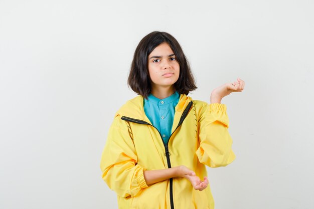 Teen dziewczyna robi gest powitalny w żółtej kurtce i patrząc niezdecydowany, widok z przodu.