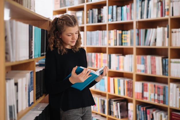 Teen dziewczyna wśród stosu książek. Młoda dziewczyna czyta książkę z półkami w tle. Otaczają ją stosy książek. Dzień książki.